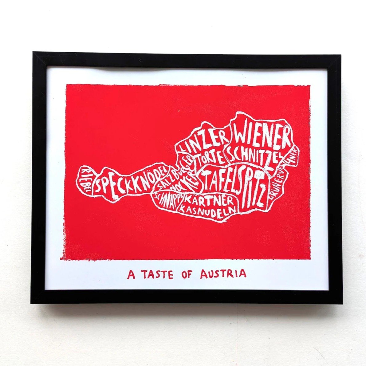 Framed art print of Austria's map with terms like "Wiener," "Schnitzel," and "Speckknödel" on a red background. Text below reads "A Taste of Austria."