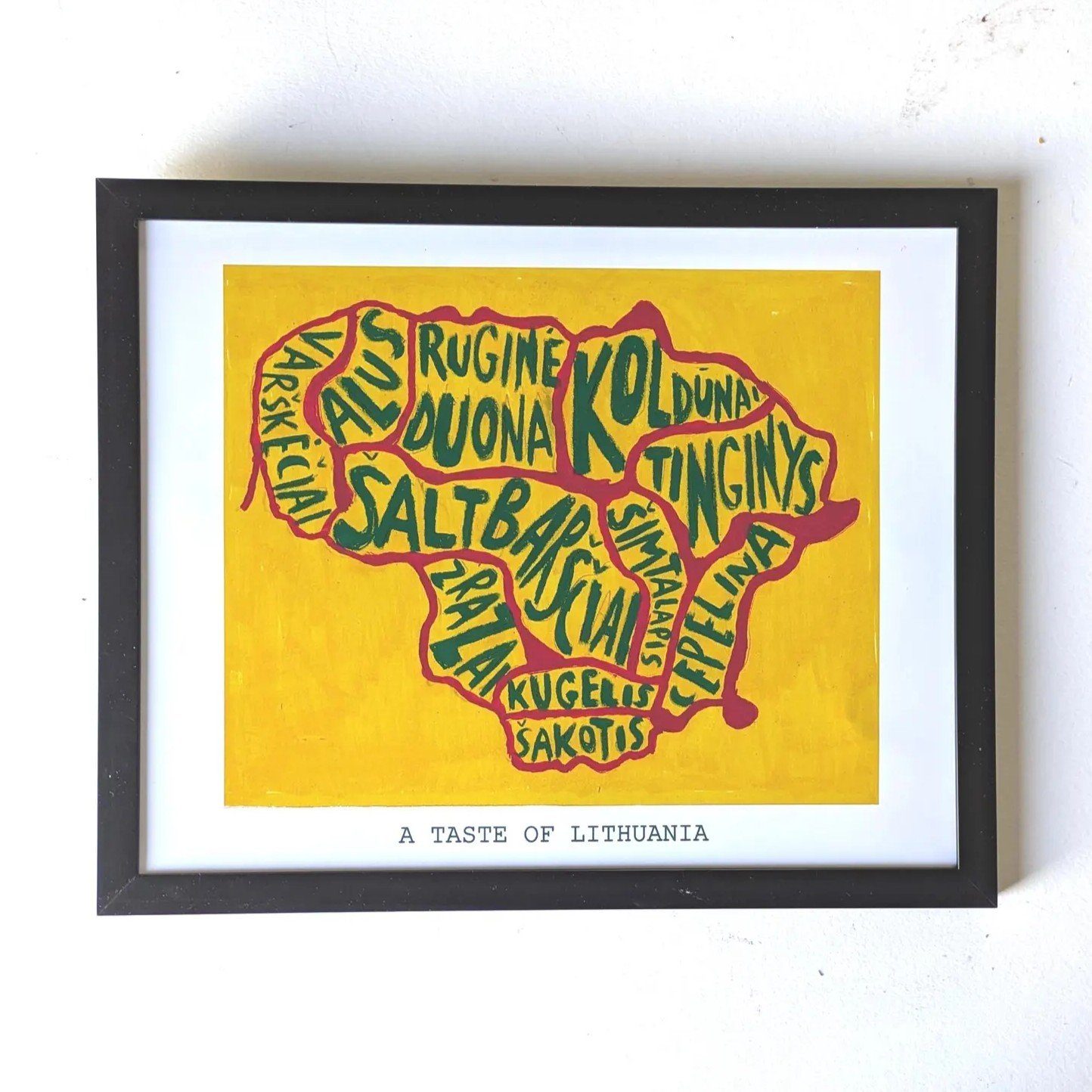 Framed art showing a map of Lithuania formed by names of traditional dishes in green text on a yellow background. Below, it says "A Taste of Lithuania."