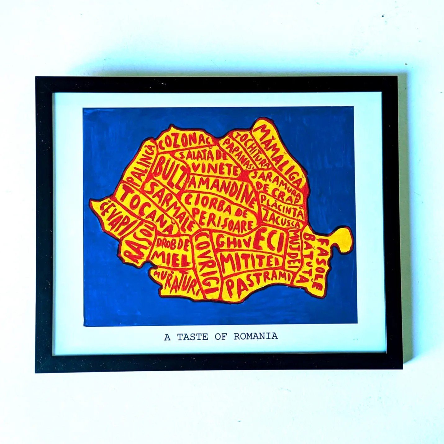Framed artwork titled "A Taste of Romania" shows a map of Romania filled with names of traditional dishes in yellow and red on a blue background.
