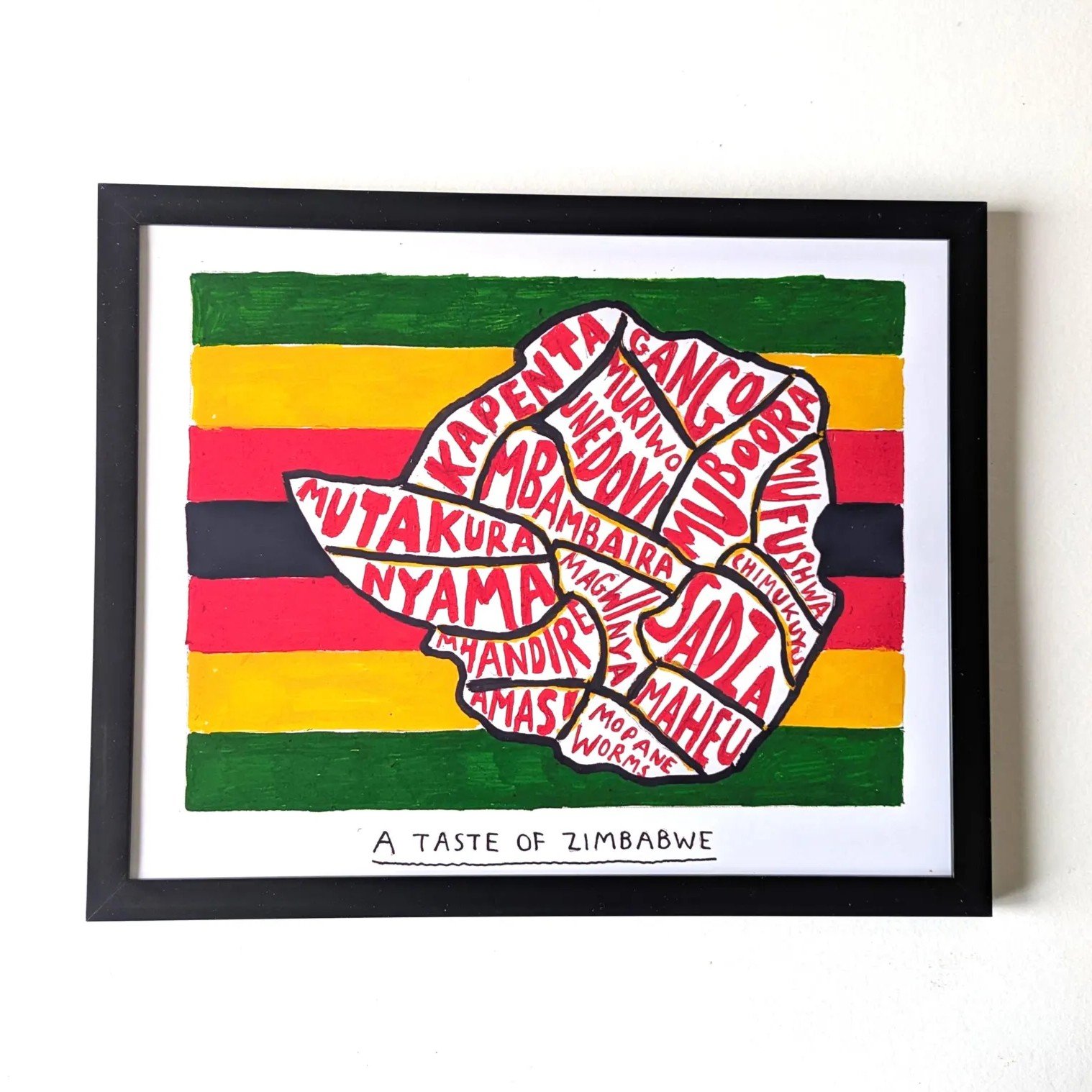 Framed artwork of Zimbabwe’s map with names of local foods in red. Background resembles Zimbabwean flag with green, yellow, red, and black stripes. Text below reads, “A Taste of Zimbabwe.”