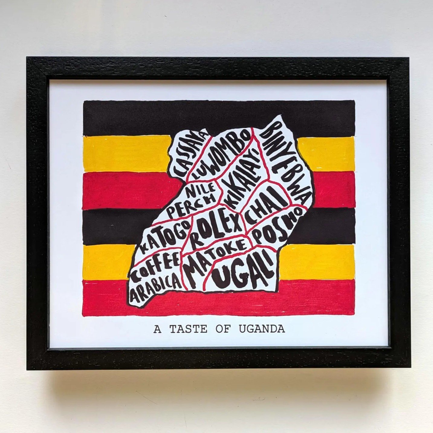 Framed artwork depicting Uganda's map filled with local food names, against Uganda's flag colors: black, yellow, and red. Caption reads "A Taste of Uganda."