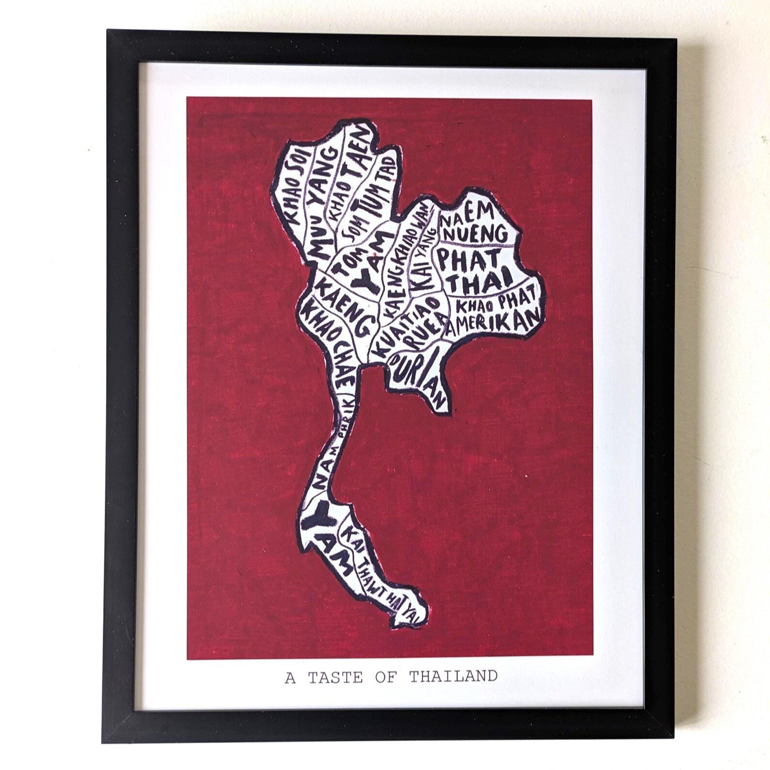 Framed artwork of Thailand's map with food names forming its shape on a red background. Text below reads "A Taste of Thailand." Bold and artistic.